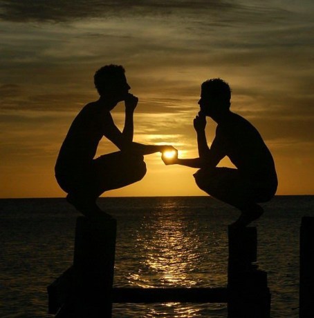 Two people holding the sun as it sets over a sea